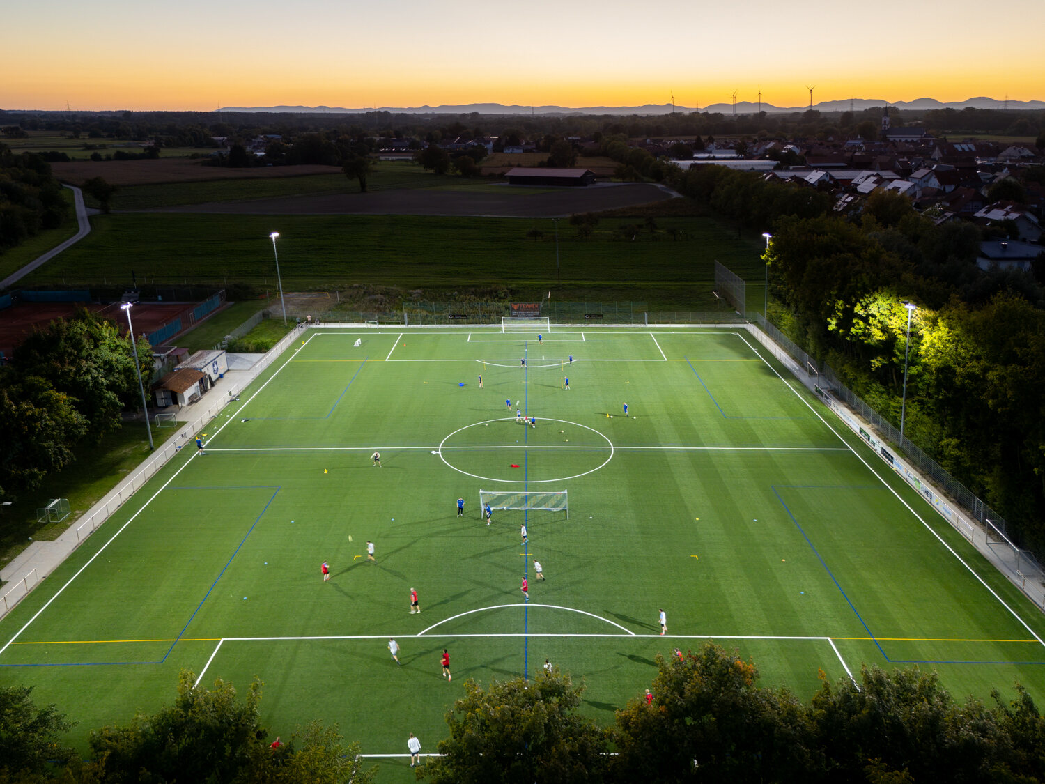 Sportplatz aus der Luft bei Sonnenuntergang, Flutlicht an, Spieler auf dem Platz